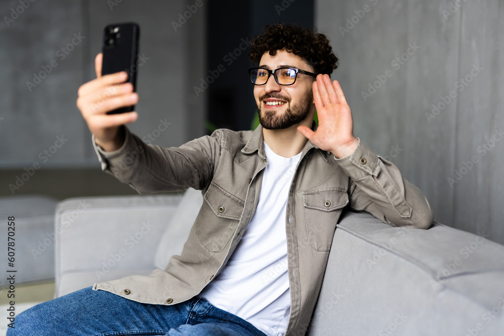 Fototapeta premium Online video call. Positive guy waving hand to webcam, having pleasant talk with friends or family. Man smiling and enjoying distant communication, sitting on floor leaning on sofa