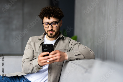 Εκτύπωση καμβά Confident bearded man is sitting on couch and typing on smartphone