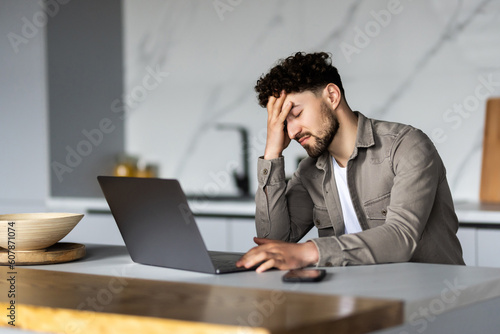 Young man in glasses sit at table looks at laptop screen touch forehead feels confused has business or pc problems