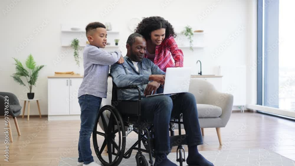Cheerful multiracial man using wheelchair holding laptop on knees while pretty woman and funny boy standing behind. Loving family of three choosing movie for watching together on Sunday at home. Cheerful multiracial man using wheelchair holding laptop on knees while pretty woman and funny boy standing behind. Loving family of three choosing movie for watching together on Sunday at home.