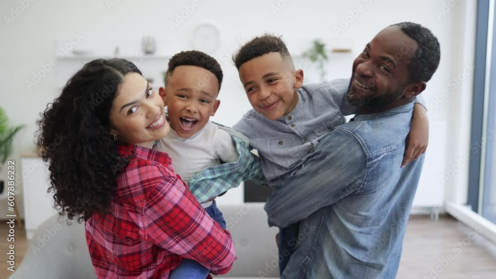 Portrait of smiling multiethnic man and woman with cute kids in arms looking over shoulder in apartment interior. Delighted parents and cute sons displaying affection for each member of loving family. Portrait of smiling multiethnic man and woman with cute kids in arms looking over shoulder in apartment interior. Delighted parents and cute sons displaying affection for each member of loving family.