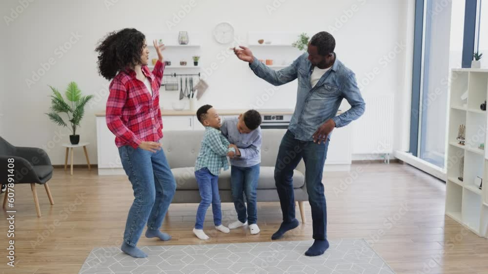 Dynamic multiethnic family of four having dancing party in bright living room of modern apartment. Healthy young parents wearing jeans teaching sons different styles of movements at leisure time. Dynamic multiethnic family of four having dancing party in bright living room of modern apartment. Healthy young parents wearing jeans teaching sons different styles of movements at leisure time.