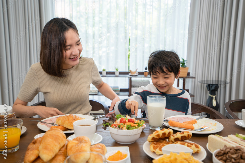Front view and selective focus of a smiling young Asian woman and a little cute boy, sitting at a table full of various breakfasts, happily teasing each other while having breakfast together at home.