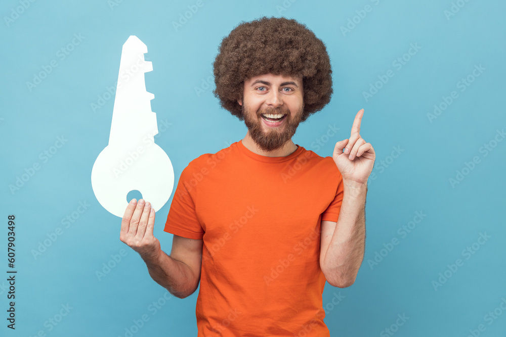 Portrait of smiling man with Afro hairstyle wearing orange T-shirt holding big paper key and pointing finger up, having idea for renting new flat. Indoor studio shot isolated on blue background. Portrait of smiling man with Afro hairstyle wearing orange T-shirt holding big paper key and pointing finger up, having idea for renting new flat. Indoor studio shot isolated on blue background.