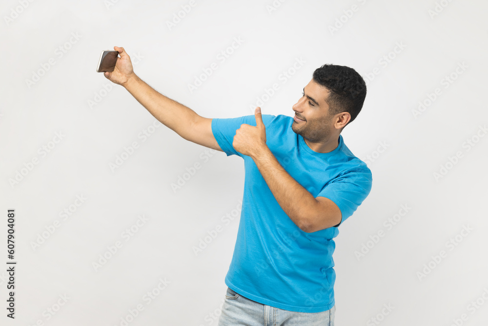 Portrait of joyful satisfied delighted man blogger in blue T- shirt holding mobile phone, has video call or livestream, showing thumb up, like gesture. Indoor studio shot isolated on gray background. Portrait of joyful satisfied delighted man blogger in blue T- shirt holding mobile phone, has video call or livestream, showing thumb up, like gesture. Indoor studio shot isolated on gray background.