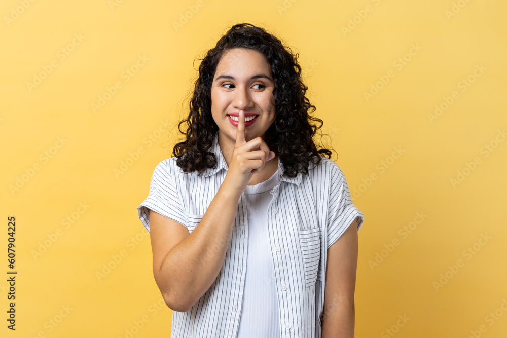 Portrait of attractive woman with dark wavy hair standing, showing hush sign and looking away with toothy smile, sharing secret. Indoor studio shot isolated on yellow background. Portrait of attractive woman with dark wavy hair standing, showing hush sign and looking away with toothy smile, sharing secret. Indoor studio shot isolated on yellow background.