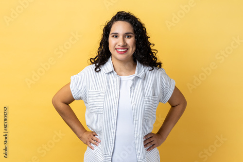 Photography Portrait of friendly positive woman with dark wavy hair standing with hands on hips, looking at camera with satisfied facial expression