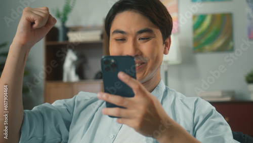 Asian man hand holding smartphone texting checking email. Excited young handsome guy using cellphone chatting with friends playing social media surprised received good news sit on sofa in living room.