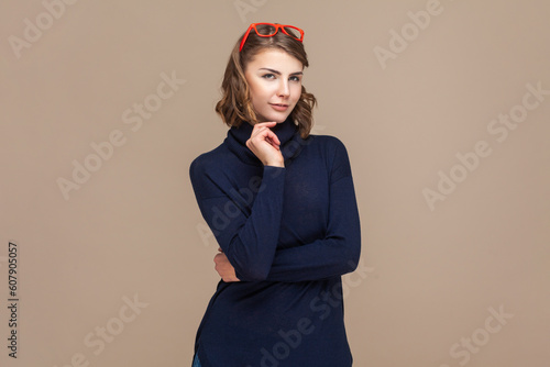 Portrait of self confident attractive woman with wavy hair in red red glasses on her head looking at camera, holding her chin. Indoor studio shot isolated on light brown background.