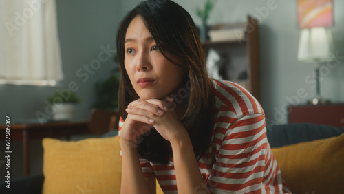 Thoughtful attractive young Asian woman props head with hand feeling bored and annoyed. Exhausted beautiful female have sad nothing to do with absent minded sit on couch at cozy home living room