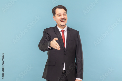 Portrait of friendly positive optimistic man with mustache standing outstretching hand, nice to meet you, wearing black suit with red tie. Indoor studio shot isolated on light blue background.