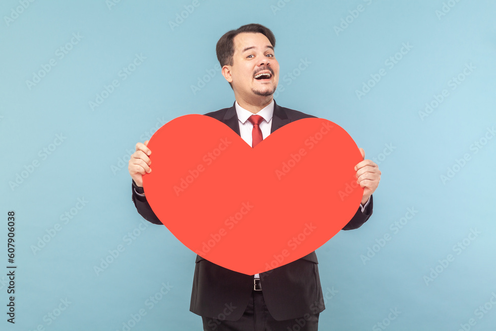 Portrait of satisfied joyful man with mustache standing holding big red card, expressing love and gentle, wearing black suit with red tie. Indoor studio shot isolated on light blue background.