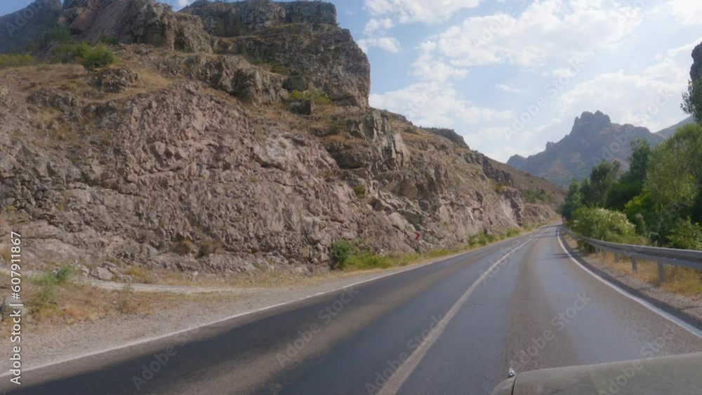 A car is driving along a road in a mountainous area. There are mountains on the side of the road, with mountains and a blue sky in the background. The road has white markings. View from the car A car is driving along a road in a mountainous area. There are mountains on the side of the road, with mountains and a blue sky in the background. The road has white markings. View from the car