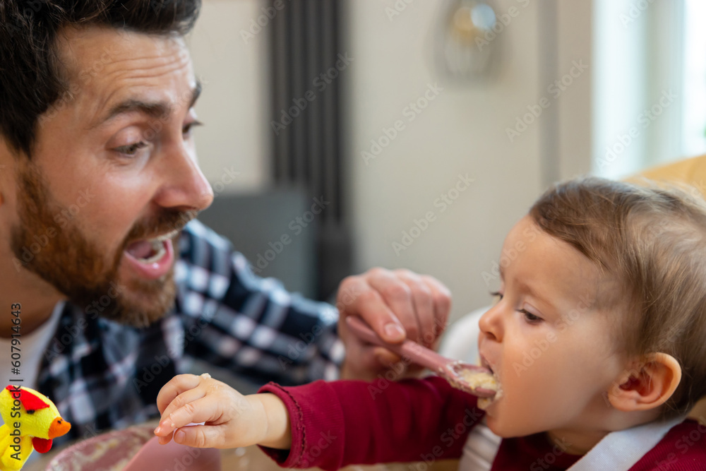 Father feeding his infant baby in home interior teaching toddler kid eating from spoon. Father feeding his infant baby in home interior teaching toddler kid eating from spoon.