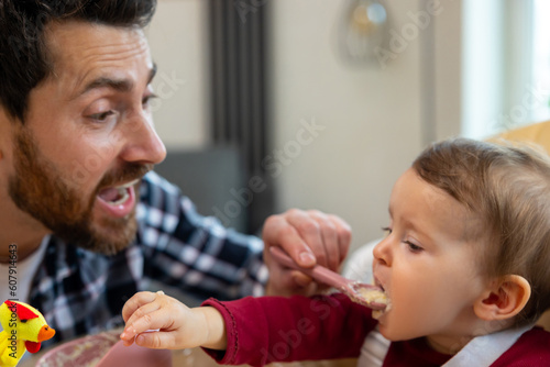 Father feeding his infant baby in home interior teaching toddler kid eating from spoon.
