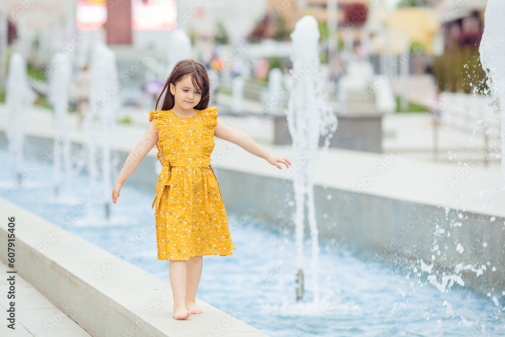 A pretty little girl in yellow dress walking barefoot near the fountain in the summer. The child has a rest near a fountain during a heat wave. A pretty little girl in yellow dress walking barefoot near the fountain in the summer. The child has a rest near a fountain during a heat wave.