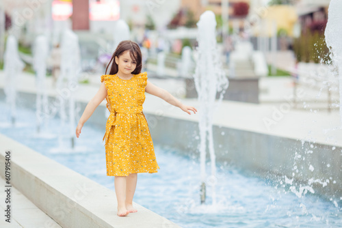 A pretty little girl in yellow dress walking barefoot near the fountain in the summer. The child has a rest near a fountain during a heat wave.