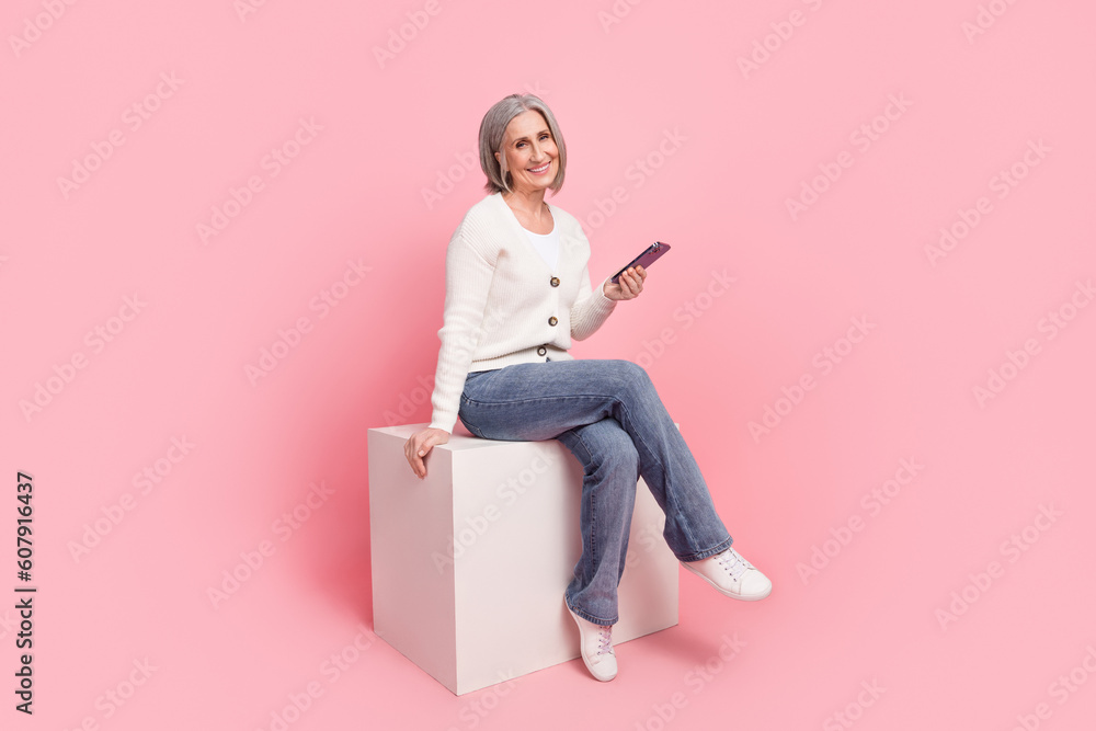 Full body photo of gorgeous mature grandma sitting cube hold telephone dressed stylish white outfit isolated on pink color background Full body photo of gorgeous mature grandma sitting cube hold telephone dressed stylish white outfit isolated on pink color background