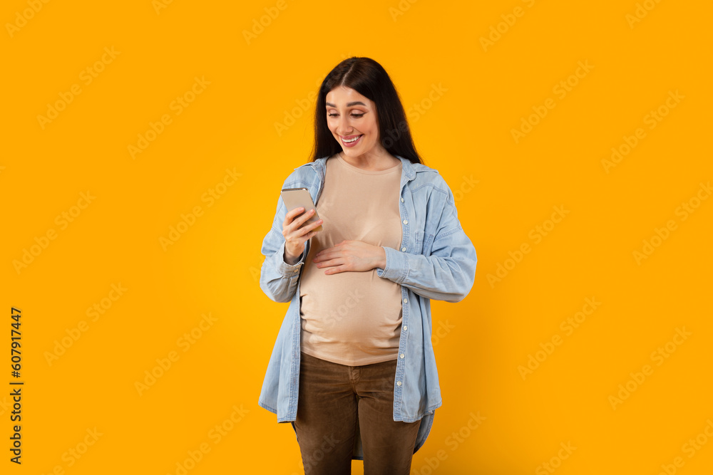 Smiling young pregnant woman using pregnancy tracker app or reading maternity blog online on phone on yellow background Smiling young pregnant woman using pregnancy tracker app or reading maternity blog online on phone on yellow background