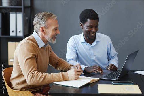 Side view portrait of handsome mature businessman using laptop with colleague at desk in office