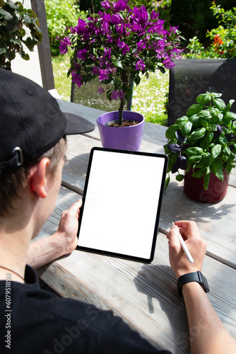 Young man using Ipad with pencil, blank empty mockup screen. Man working in the garden. Freelance, student lifestyle, drawing, gardening,