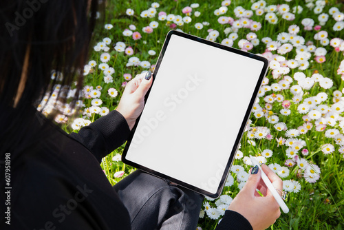 Young woman using Ipad with pencil, blank empty mockup screen. in a field of flowers. Women are drawing in the garden: Freelance, student lifestyle, drawing, gardening.
