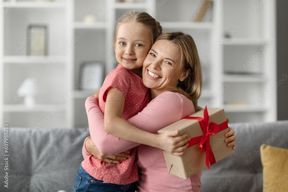 Cute little girl holding gift box and hugging her mom at home Cute little girl holding gift box and hugging her mom at home