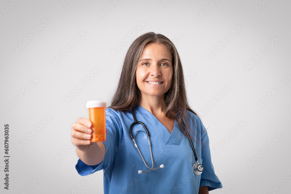 Cheerful senior woman doctor in uniform showing jar of pills to camera, posing on grey studio background Cheerful senior woman doctor in uniform showing jar of pills to camera, posing on grey studio background
