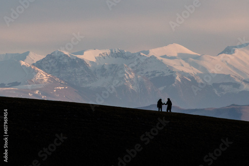 A very distant couple shaking hands on top of a mountain with distant hills and mountains covered by snow © Massimo