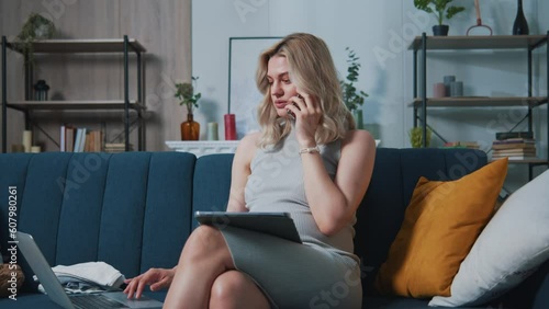 Pregnant beautiful caucasian woman talking by smartphone, using a notebook and tablet while sitting on a blue sofa. Young future mother having tedious phone talk, sighing and rolling eyes
