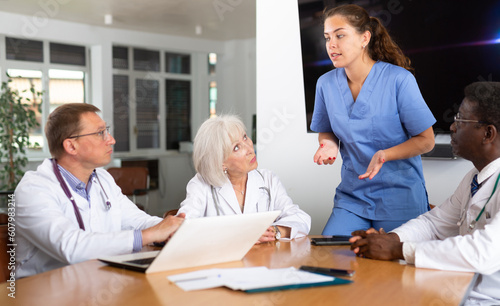 Group of different people in medical uniforms discussing issues while sitting at table in office