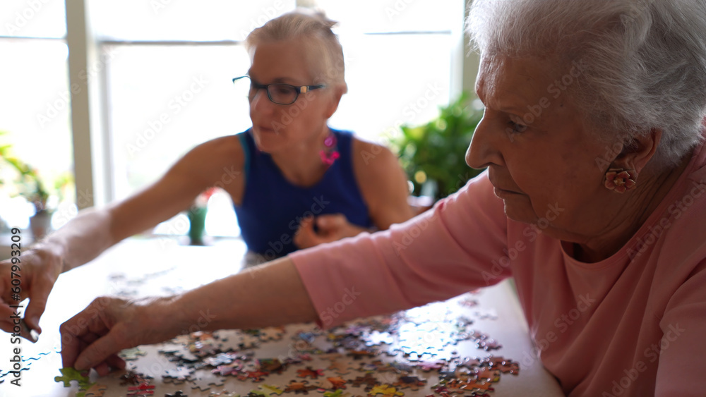Naklejka premium Closeup of happy pretty elderly woman organizing puzzle pieces while daughter moves pieces to fit together. Concept of spending time with loved ones.