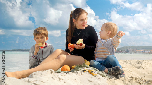 A happy woman enjoys a beach picnic with her two sons, eating fresh fruits and feeling joy. Family vacation and weekend on nature.