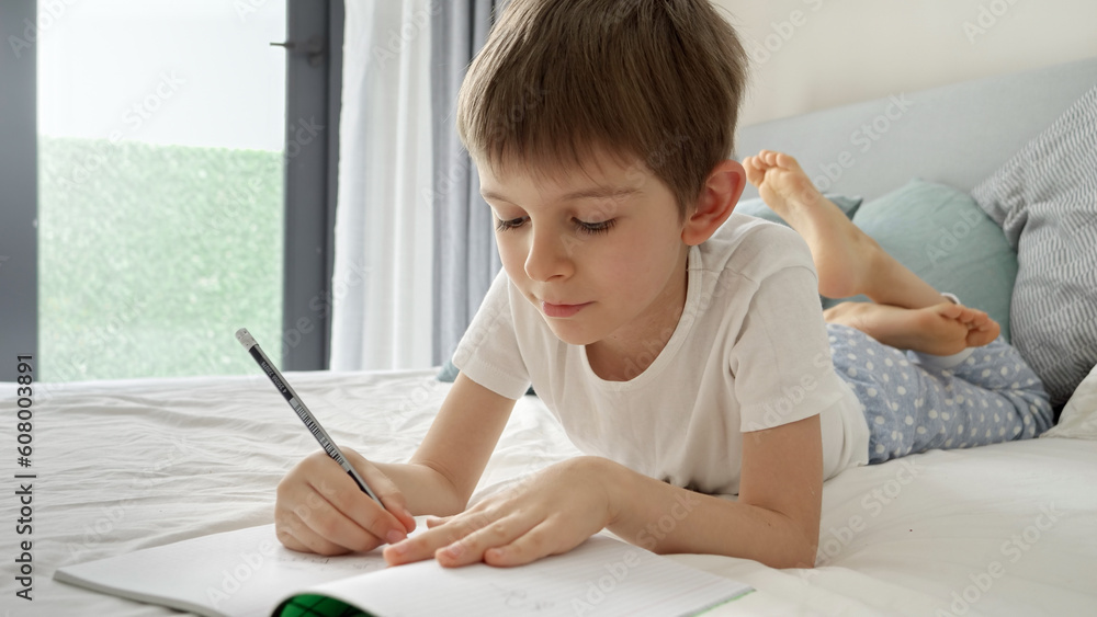 Portrai of little boyworking on his homework in bed, writing in a notebook with concentration. Importance of education and child development in a remote learning environment. Portrai of little boyworking on his homework in bed, writing in a notebook with concentration. Importance of education and child development in a remote learning environment.