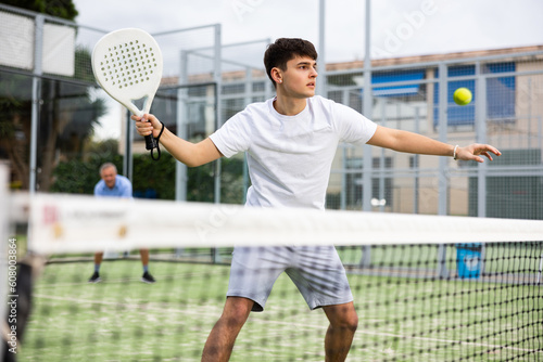 Portrait of emotional determined young guy playing padel tennis on open court in summer, swinging racket to return ball over net. Sportsman ready to hit volley