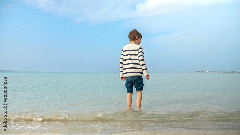 Rear view of little boy in sweater walking on sandy beach and entering sea water. Holiday, vacation, weekend at nature.