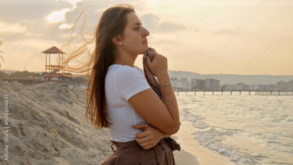 Portrait of young worried woman standing at the beach and looking on sea waves rolling on shore.