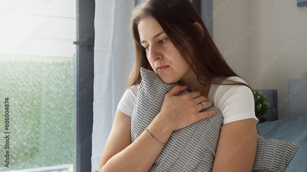 With tears in her eyes, the young woman sits on the bed, her arms wrapped around a cushion in a tight embrace. The concept of emotional distress, loneliness, and heartbreak. With tears in her eyes, the young woman sits on the bed, her arms wrapped around a cushion in a tight embrace. The concept of emotional distress, loneliness, and heartbreak.