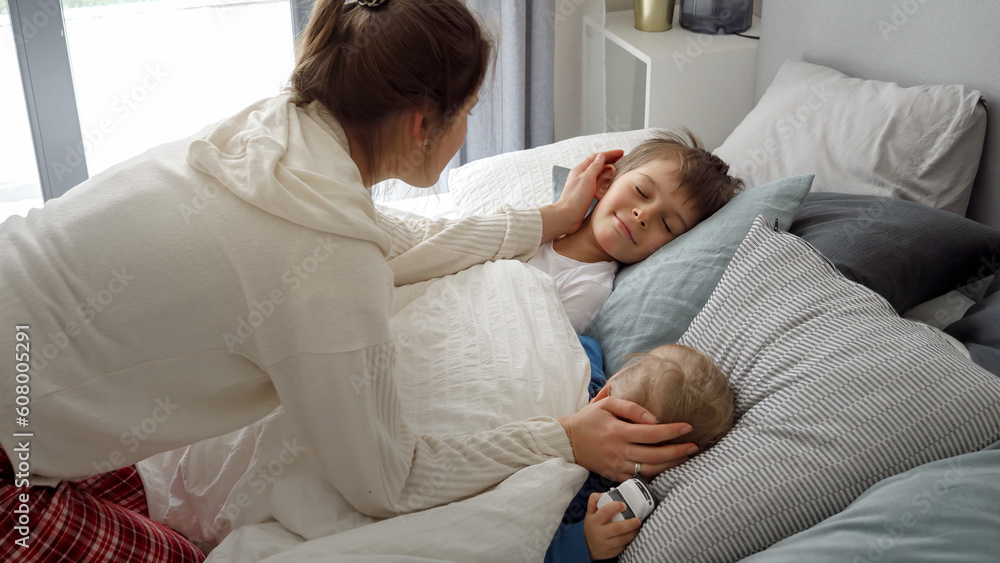 Portrait of smiling loving mother covering with blanket and looking at her children sleeping in bed. Concept of family happiness, relaxing at home, having fun in bed, parent and cheerful kids Portrait of smiling loving mother covering with blanket and looking at her children sleeping in bed. Concept of family happiness, relaxing at home, having fun in bed, parent and cheerful kids