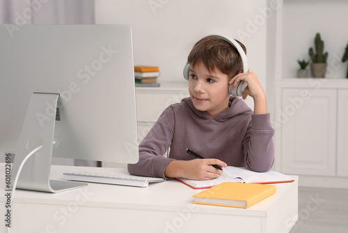 Boy writing in notepad while using computer and headphones at desk in room. Home workplace