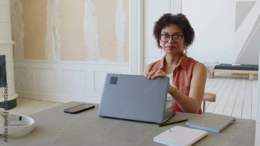 Medium portrait shot of young biracial woman in glasses and sleeveless shirt working on laptop in trendy contemporary apartment, then closing computer, looking at camera and smiling Medium portrait shot of young biracial woman in glasses and sleeveless shirt working on laptop in trendy contemporary apartment, then closing computer, looking at camera and smiling