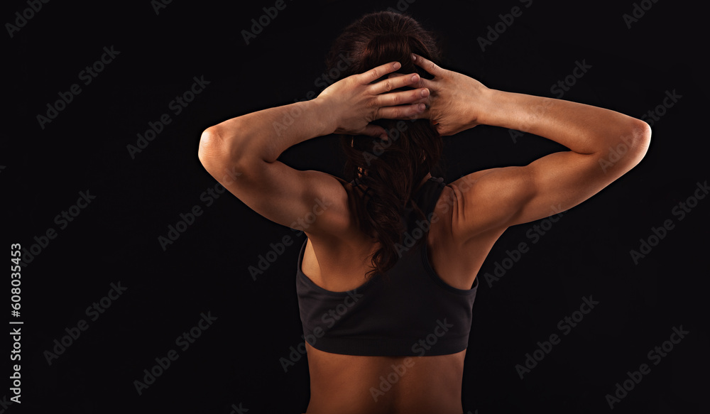 Naklejka premium Female sporty muscular with ponytail doing stretching workout of the shoulders, blades in sport bra, holding the neck hands on black background with empty copy space. Back view.