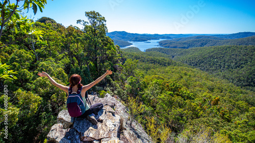 Beautiful girl sits on a rock shelf enjoying view of the Advancetown lake after successfully climbing the Pages Pinnacle. Hidden gems in Springbrook National Park, Gold Coast, Queensland, Australia