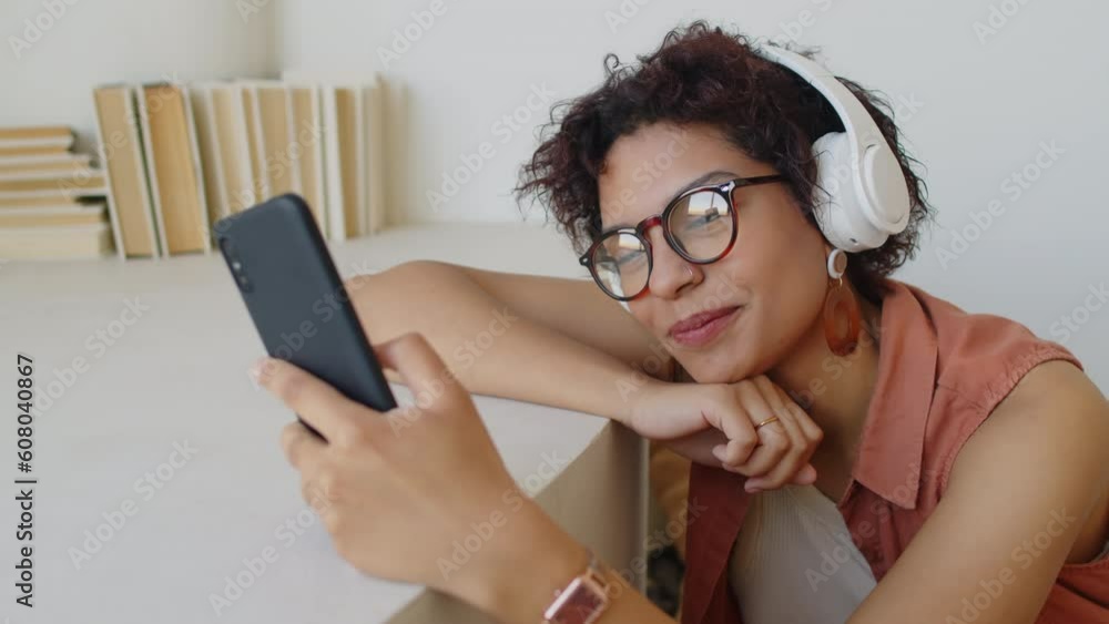 Medium close-up shot of young biracial woman with auburn curly hair, in glasses and large white wireless headphones, sitting with arm under chin, scrolling through messages on smartphone and smiling Medium close-up shot of young biracial woman with auburn curly hair, in glasses and large white wireless headphones, sitting with arm under chin, scrolling through messages on smartphone and smiling