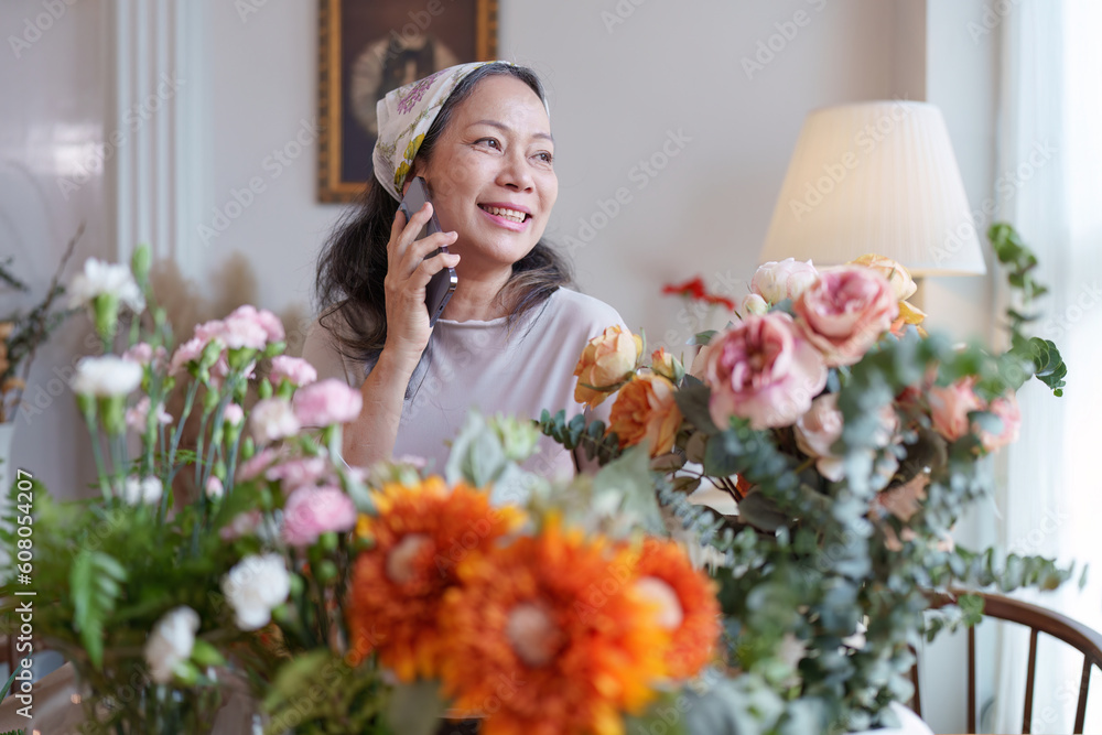 Startup, small business, flower shop. Older florist putting a flower on a vase and talking with customer by phone