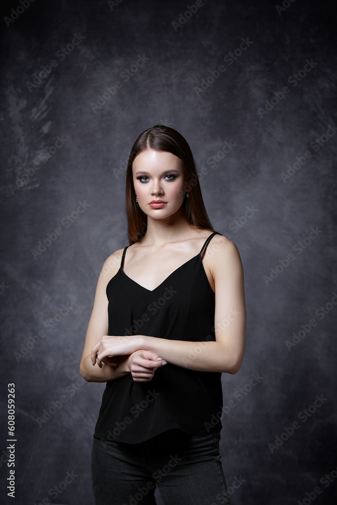 Portrait of female in black tank top with straps and jeans. Woman with earrings in the ear on gray background. Portrait of female in black tank top with straps and jeans. Woman with earrings in the ear on gray background.