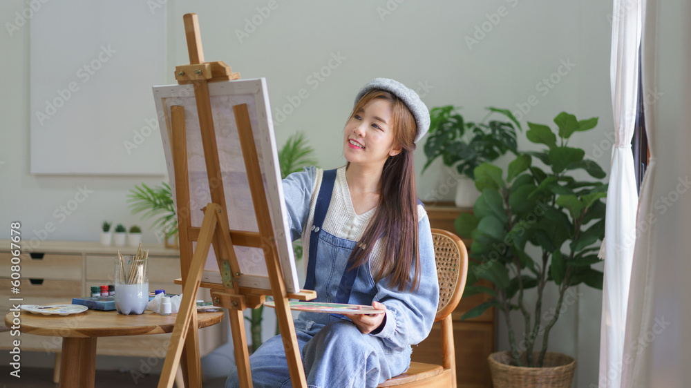 Young female artist holds color palette and use brush to painting artwork on canvas in home studio