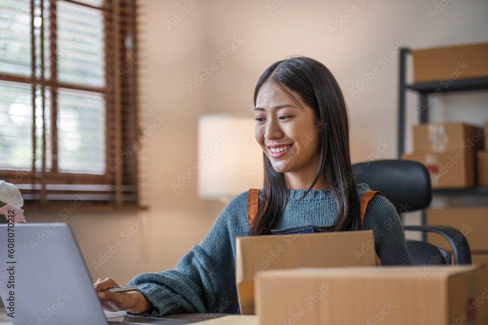 Happy young asian woman startup small business freelance holding parcel box and computer laptop and sitting on chair, Online marketing packing box delivery concept Happy young asian woman startup small business freelance holding parcel box and computer laptop and sitting on chair, Online marketing packing box delivery concept