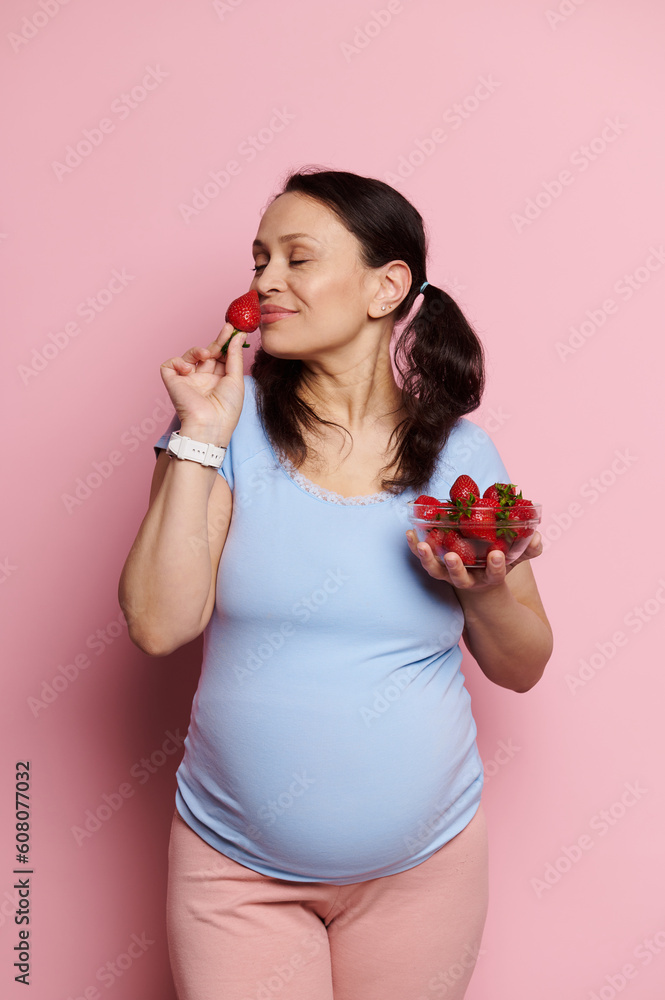 Beautiful glamorous pregnant woman in a blue t-shirt and pink pants, sniffing fresh ripe strawberries, enjoying a healthy diet during pregnancy, posing with bowl of berries on isolated pink background Beautiful glamorous pregnant woman in a blue t-shirt and pink pants, sniffing fresh ripe strawberries, enjoying a healthy diet during pregnancy, posing with bowl of berries on isolated pink background