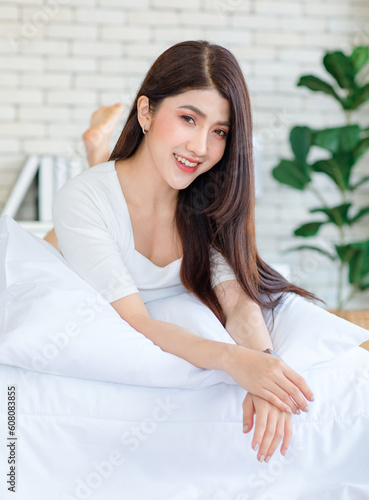 Portrait closeup shot of Millennial Asian beautiful cheerful happy female model in casual outfits with wrist watch laying lying down on white pillow posing smiling look at camera on bed in bedroom